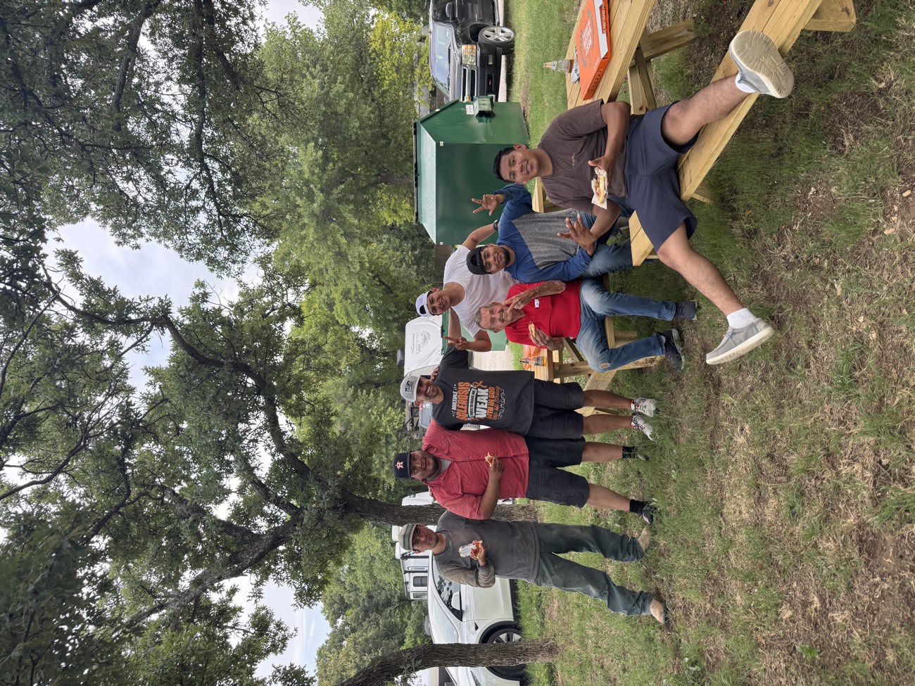 A group of smiling guests sharing pizza at a picnic table under the oak trees